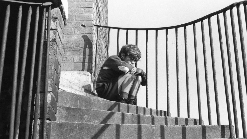 Black and white photo of a boy in shorts and torn jumper sitting on some curved steps 